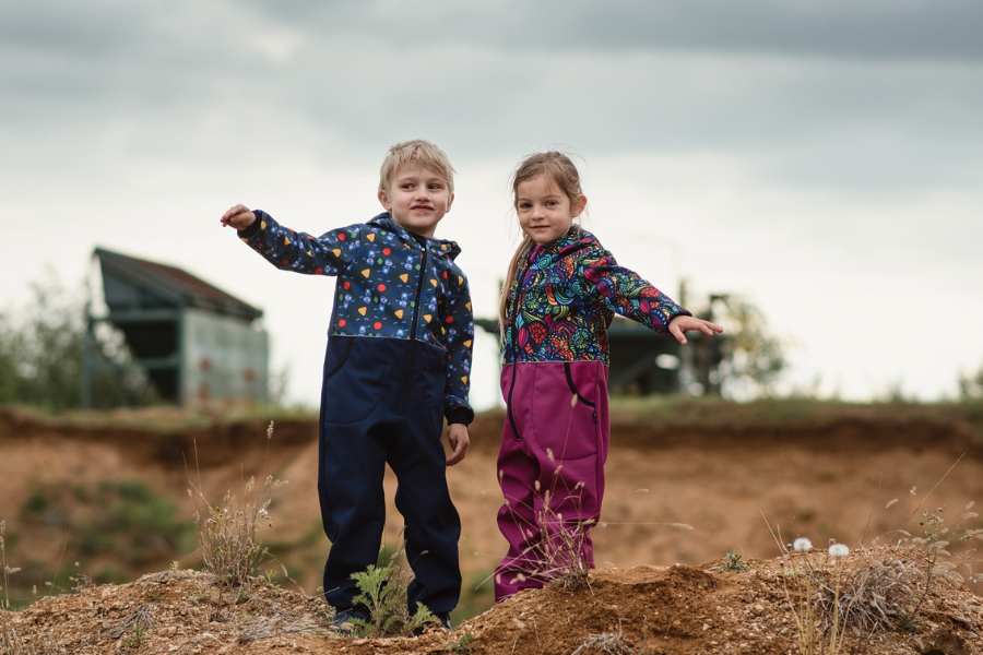 Two small children, a girl and a boy in colorful overalls are standing on a hill
